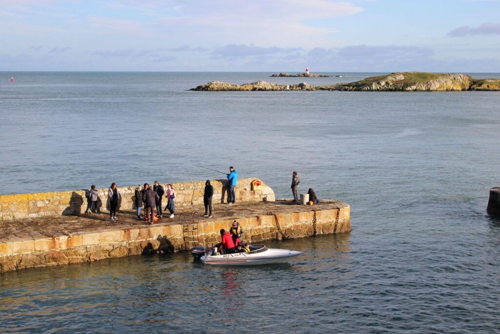 People fishing and gathering on the stone pier at Coliemore Harbour, with a small motorboat alongside and rocky islands offshore.