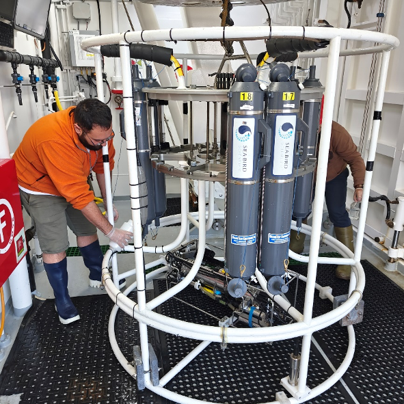 Two crew members prepare a circular water-sampling instrument fitted with sample bottles and sensors aboard a research vessel.